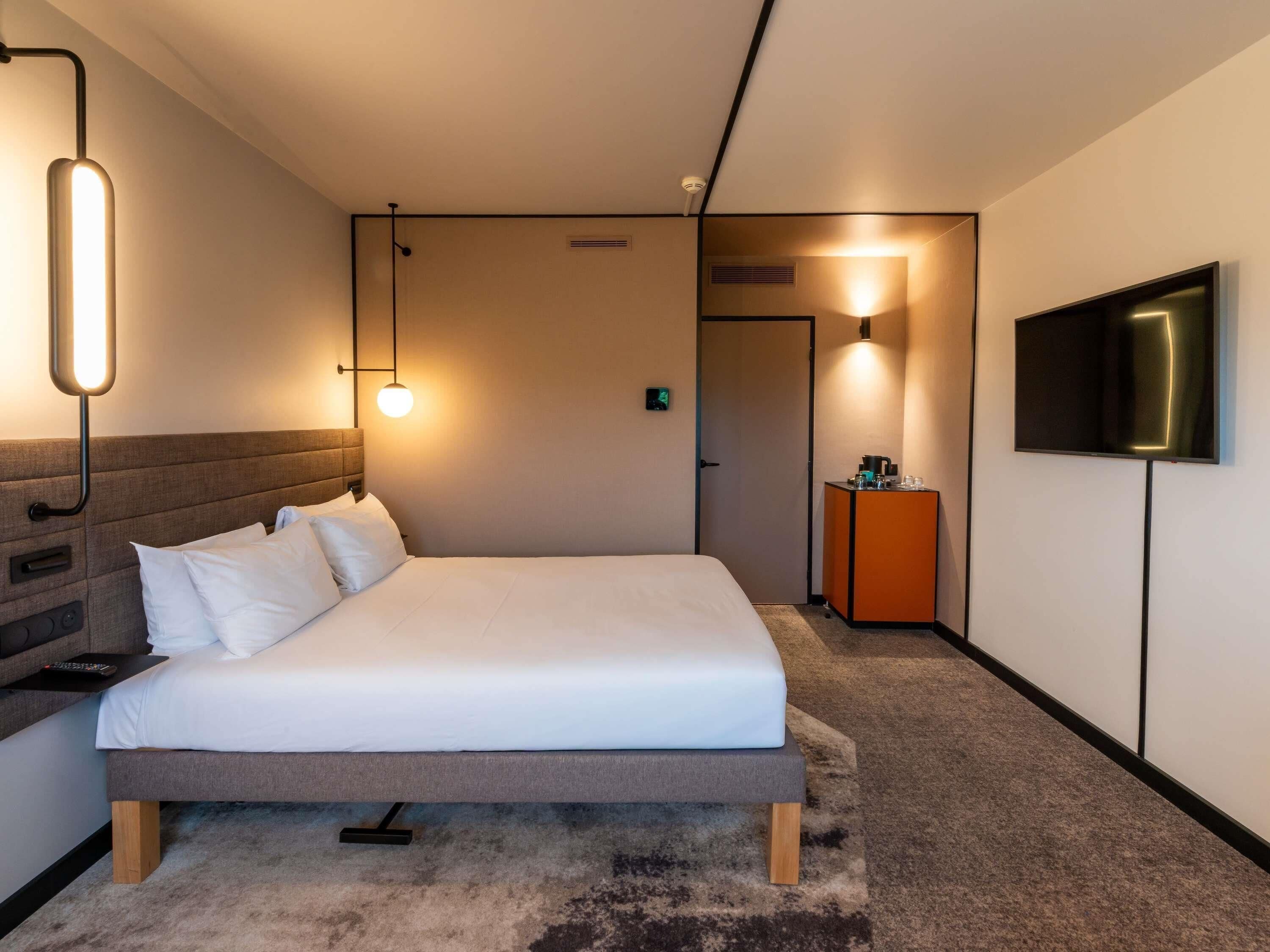 Ein modernes Hotelzimmer mit Bett, Nachttisch, Lampe, Fernseher und einer Tür zu einem weiteren Raum.