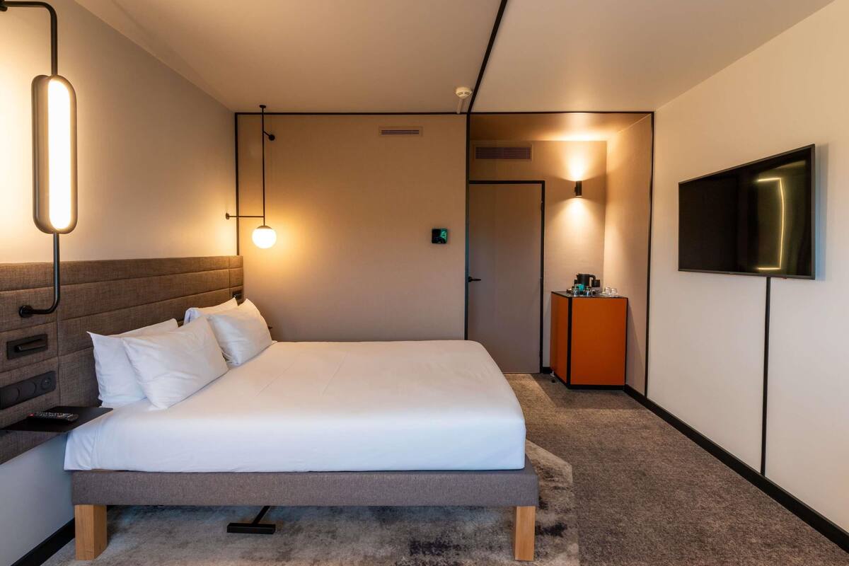 A modern hotel room with a bed, bedside table, lamp, TV, and a door leading to another room.