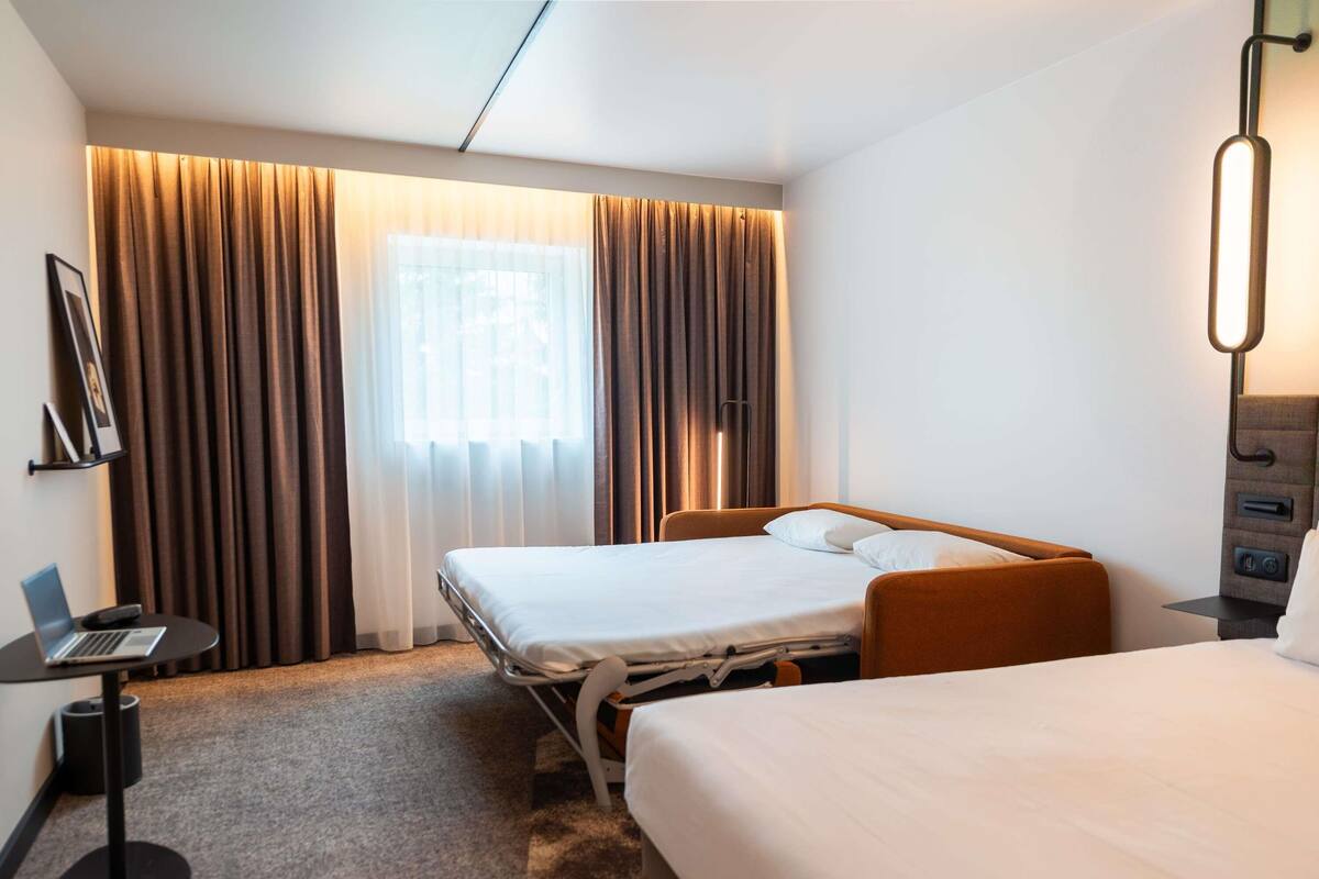A hotel room with a bed, a desk with a laptop, a bedside table, and a lamp.