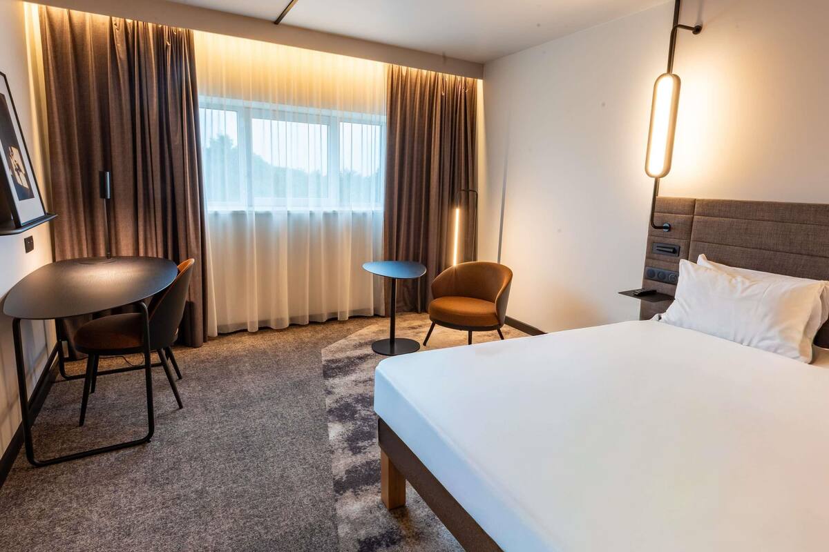 A hotel room with a bed, a desk, a chair, a lamp, and a window with curtains.