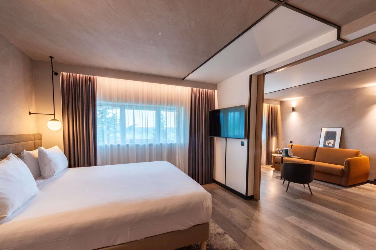 A modern hotel room with a large bed, a flat-screen TV, a sofa, and a dining area.