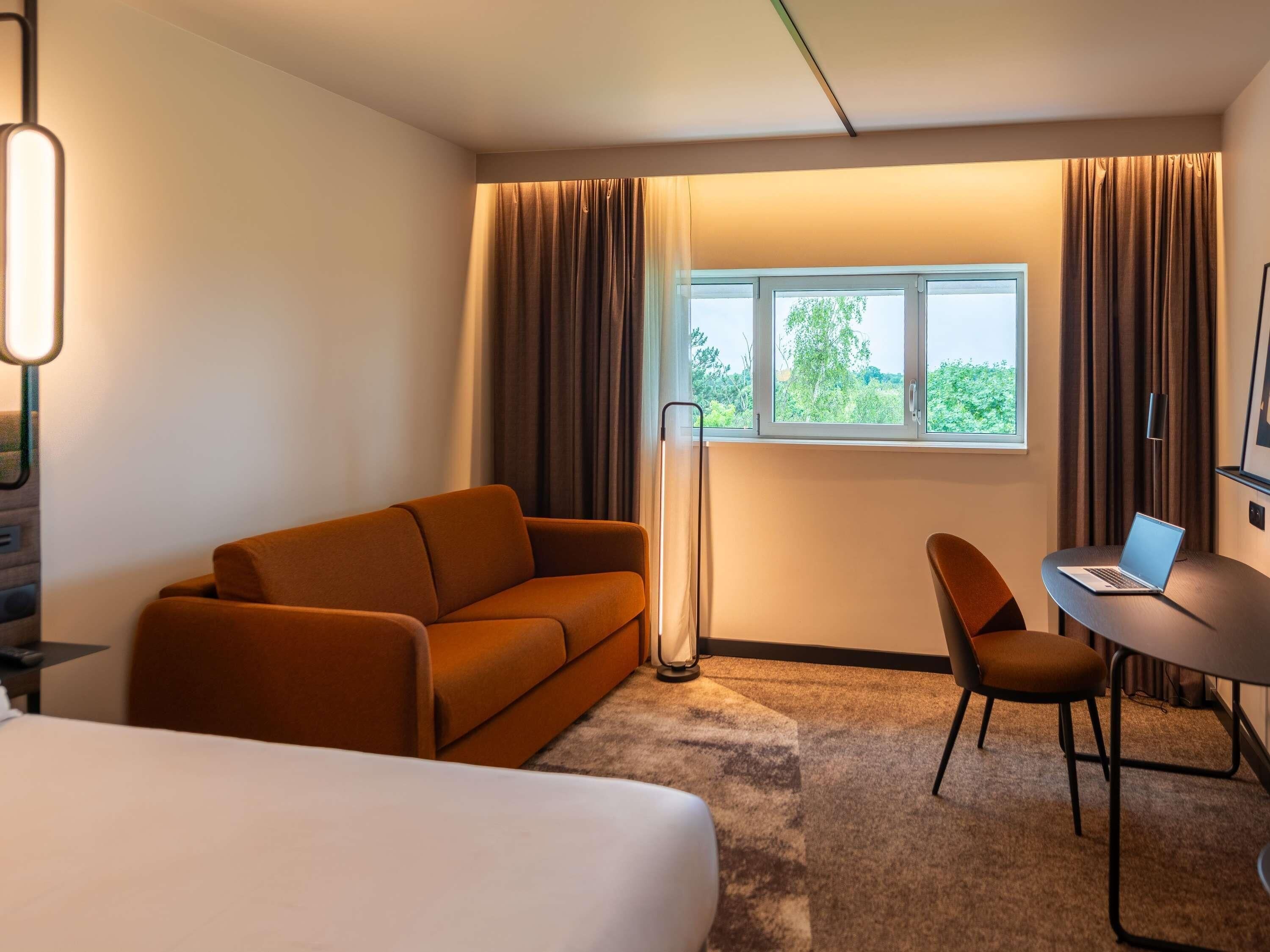 Habitación de hotel con cama, sofá, escritorio con una computadora portátil, lámpara de pie y ventana con cortinas.