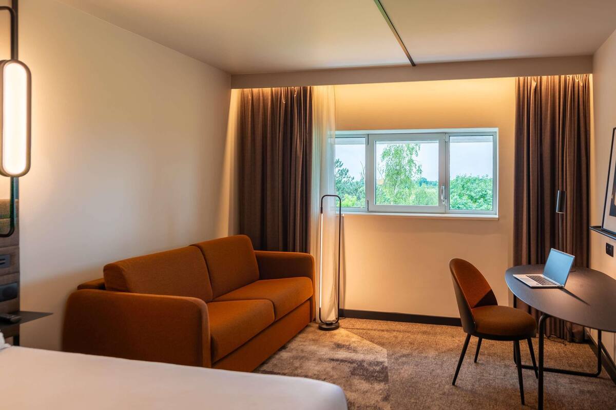 A hotel room with a bed, a sofa, a desk with a laptop, a standing floor lamp, and a window with curtains.