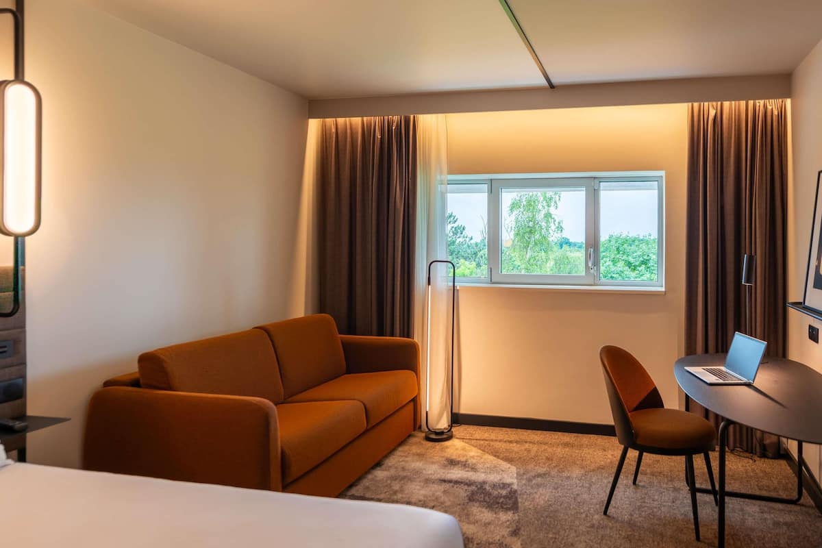 Habitación de hotel con cama, sofá, escritorio con una computadora portátil, lámpara de pie y ventana con cortinas.