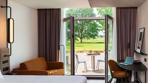 Habitación de hotel con cama, escritorio con computadora portátil, sofá y vista a un área verde a través de una puerta de vidrio.