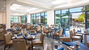 A spacious hotel dining area with multiple tables and chairs, large windows, and a view of the outdoors.