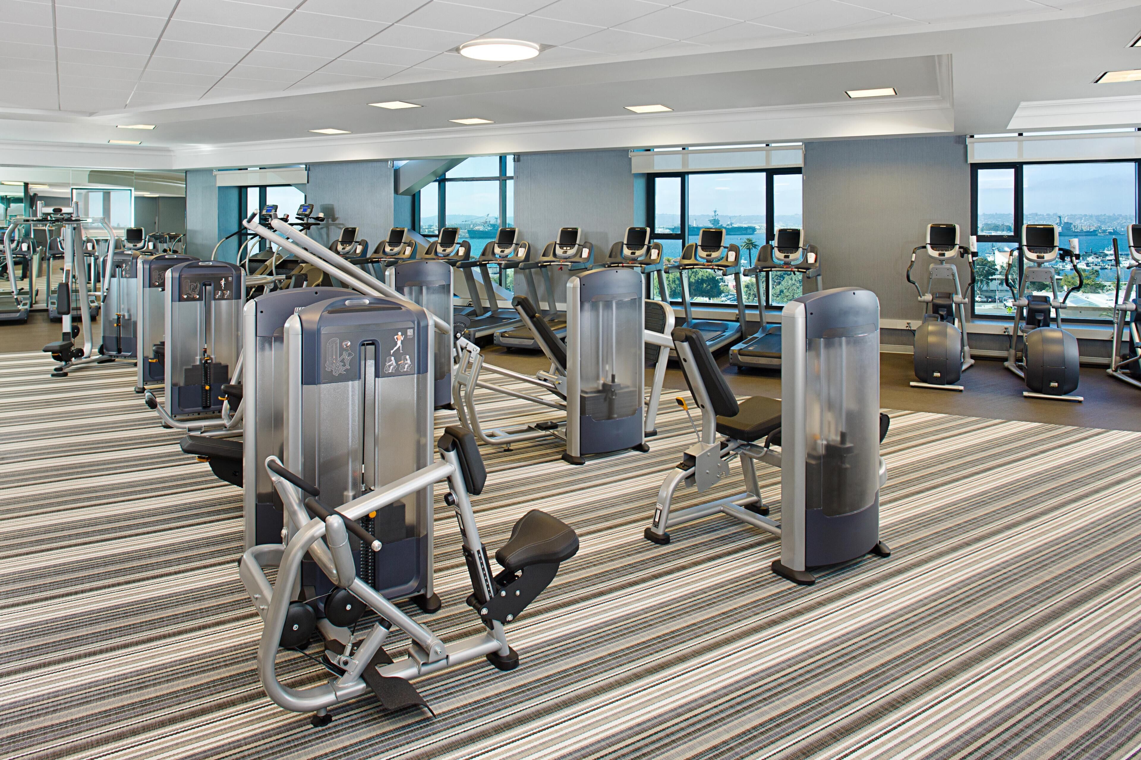 A gym with various exercise machines, large windows, and a carpeted floor.