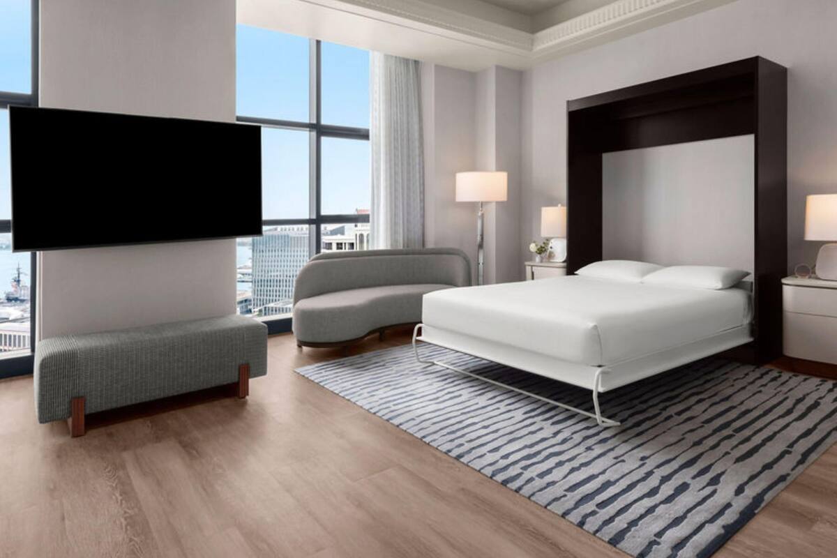 A modern hotel room with a large bed, a sofa, a TV, and a city view.