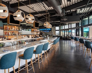 Een bar met een lange toonbank, barstoelen en uitzicht op de stad door grote ramen.