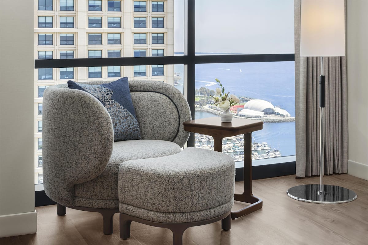 A modern room with a large window, a grey upholstered sofa with a blue pillow, a small round table with a vase, and a floor lamp.