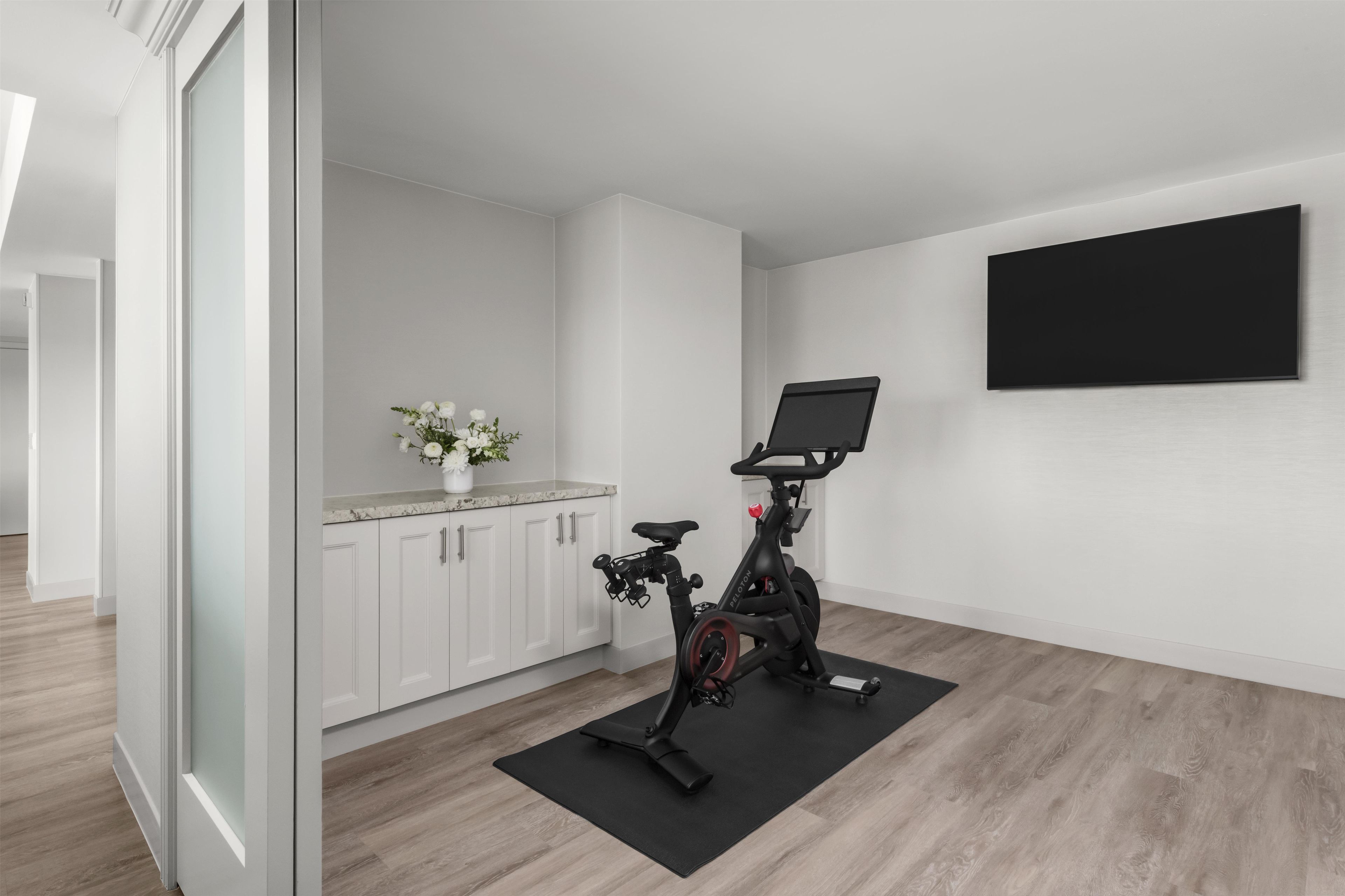 A modern room with a stationary bike, a flat-screen TV, and a kitchen area with white cabinets.