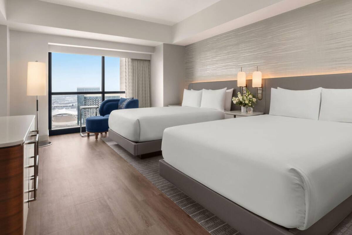 A hotel room with two beds, a desk, a chair, and a large window with a city view.