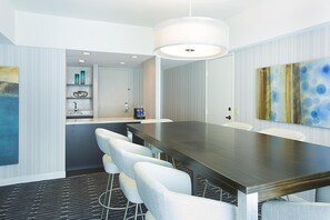 A conference room with a long table, white chairs, a modern pendant light, and a kitchen area in the background.