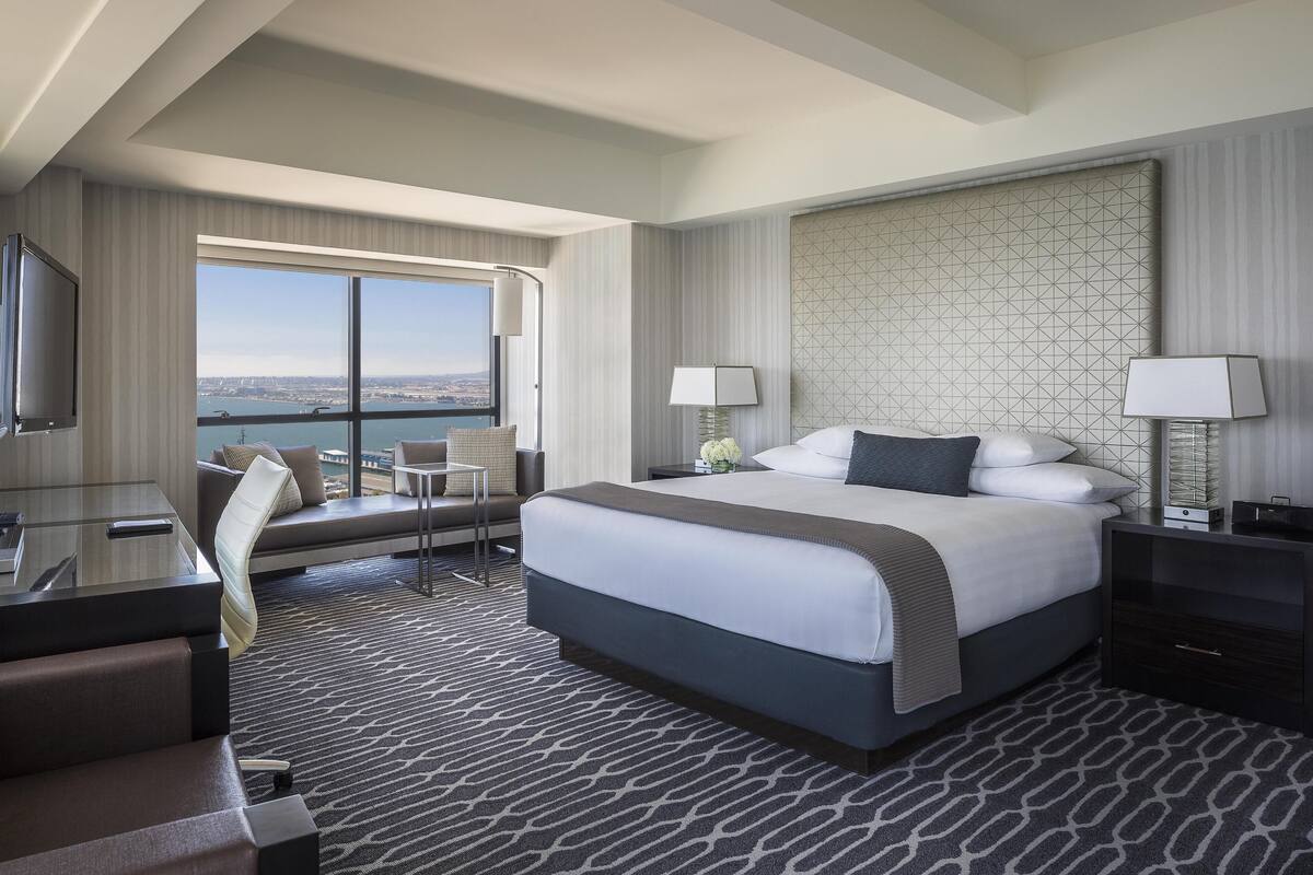 A modern hotel room with a large bed, a desk, a chair, and a view of the city.