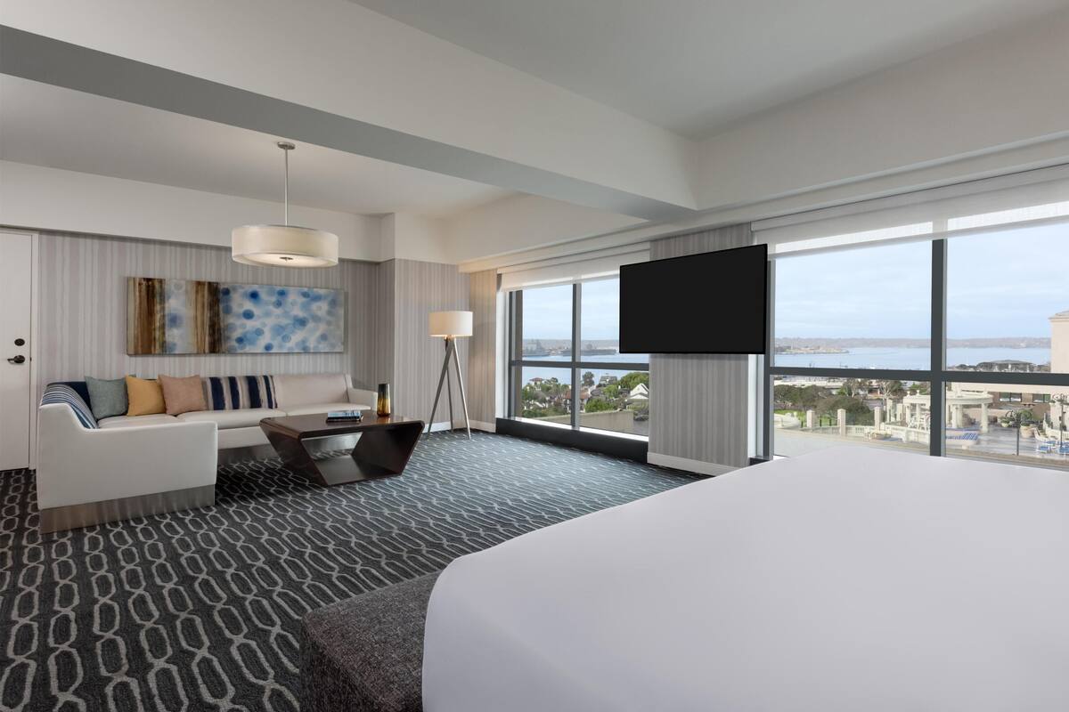 A hotel room with a large bed, a sofa, a coffee table, a TV, and a city view.