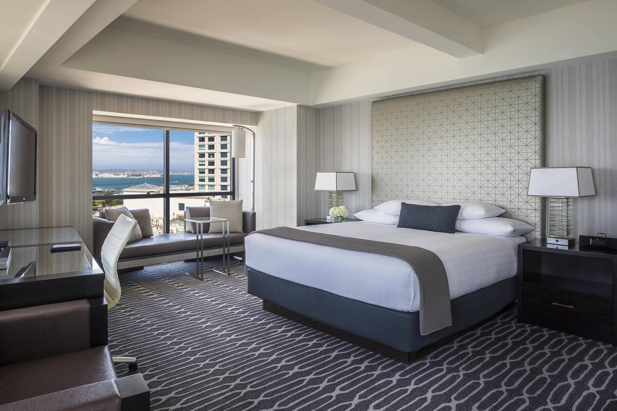 A hotel room with a large bed, a desk, a chair, a TV, and a view of the city and ocean.