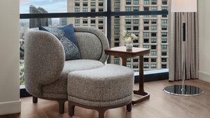 Un salon moderne avec un canapé et un pouf, une petite table sur laquelle est posé un vase de fleurs, une lampe sur pied et une vue sur le paysage urbain à travers de grandes fenêtres.