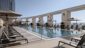 A rooftop pool with lounge chairs and umbrellas, a clear sky, and a high-rise building in the background.