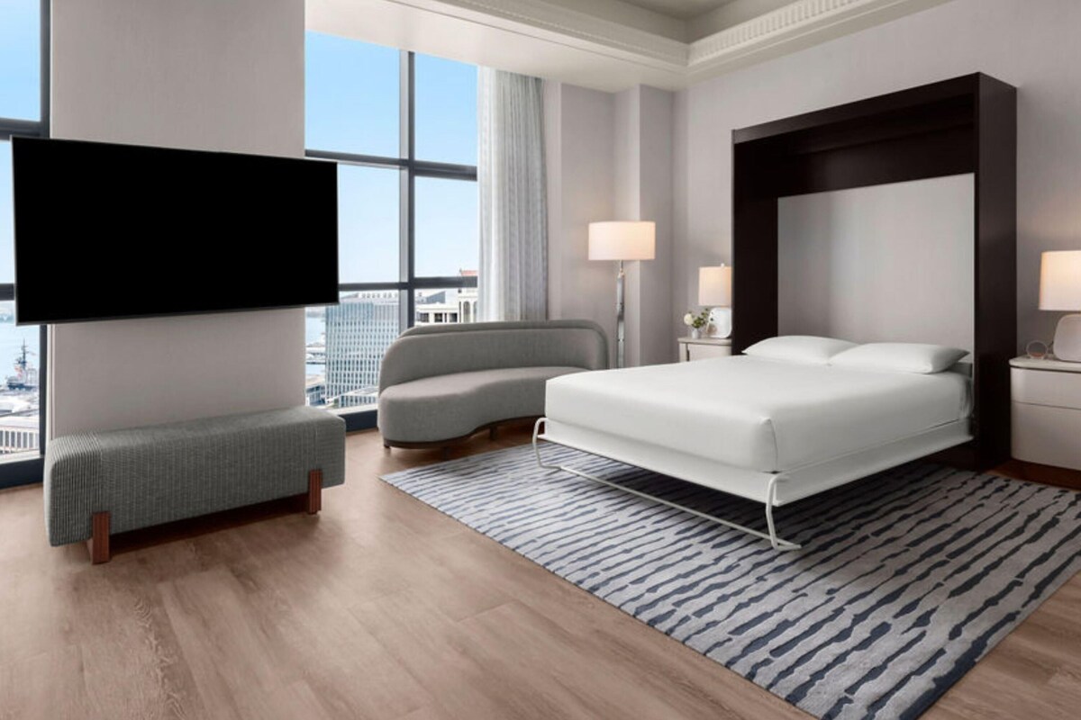 A modern hotel room with a large bed, a sofa, a TV, and a city view.