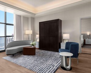 Une chambre d’hôtel moderne avec un canapé gris, un fauteuil bleu, une petite table ronde, une armoire sombre et une grande fenêtre donnant sur la ville.