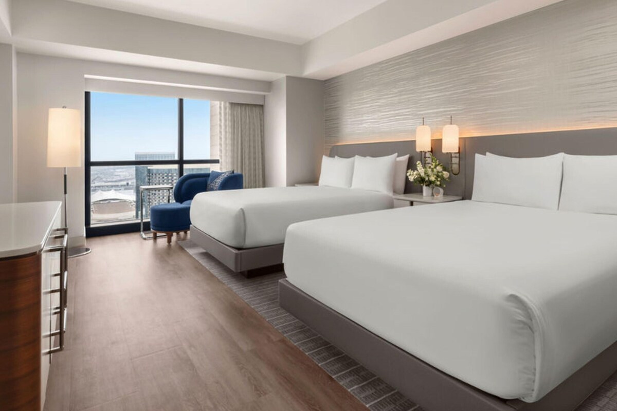 A hotel room with two beds, a desk, a chair, and a large window with a city view.