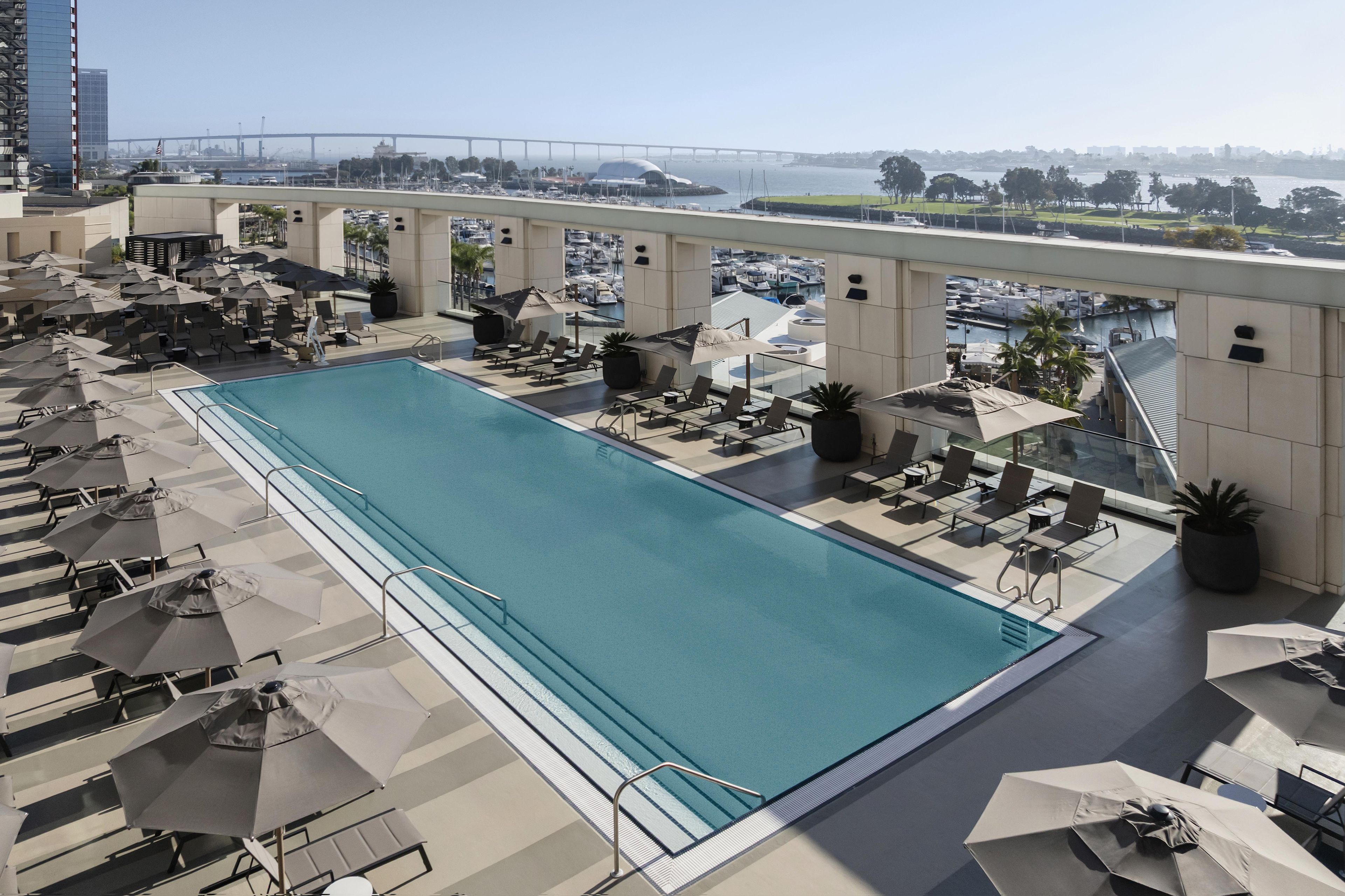 A rooftop pool with lounge chairs and umbrellas, a view of a bridge, and a marina in the background.