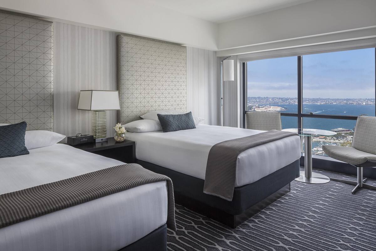 A hotel room with two beds, a chair, a small table, and a large window offering a city view.