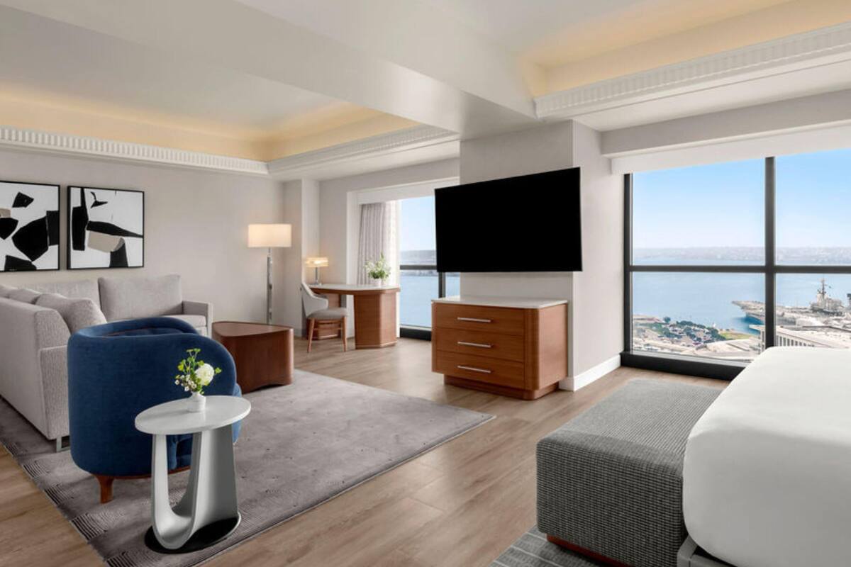 A modern hotel room with a large bed, a flat-screen TV, a desk, a sofa, and a view of the sea.