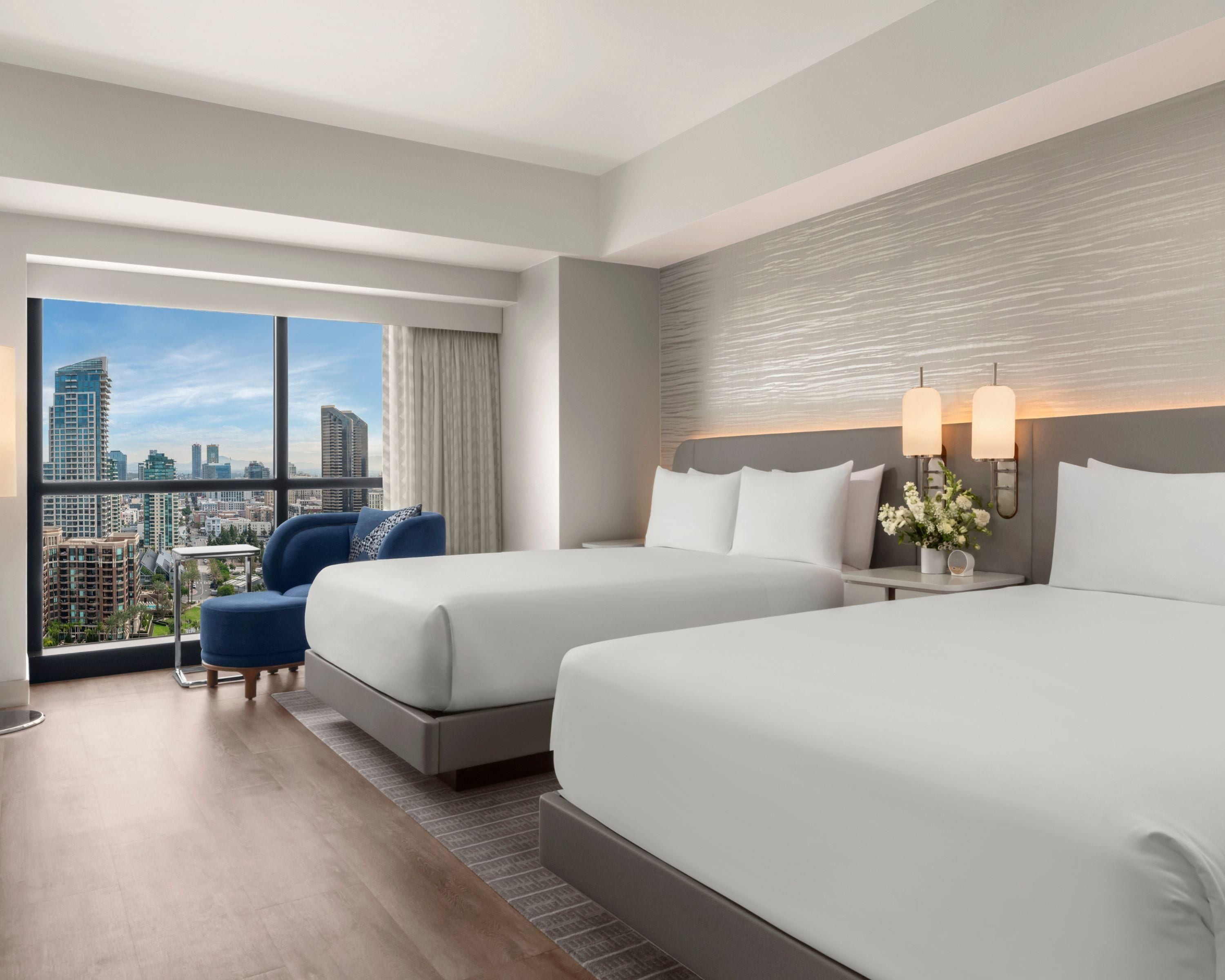 A hotel room with two beds, a sofa, a nightstand with lamps, and a view of the cityscape through large windows.