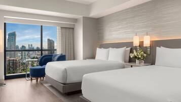 A hotel room with two beds, a sofa, a nightstand with lamps, and a view of the cityscape through large windows.