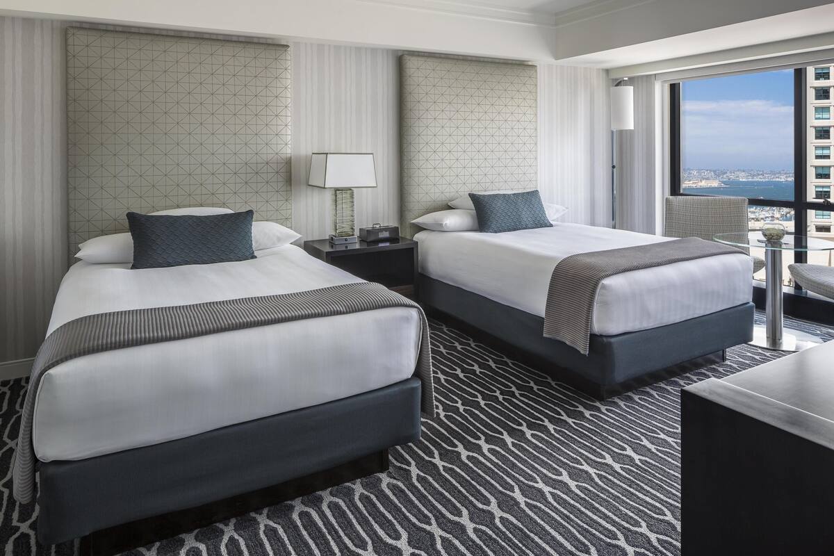 A hotel room with two beds, a nightstand, a lamp, a chair, and a view of the city.
