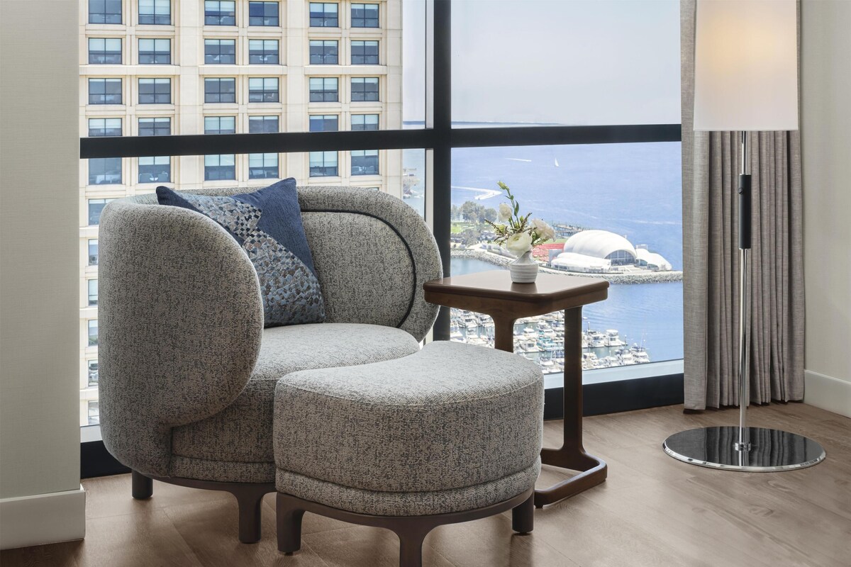 A modern room with a large window, a grey upholstered sofa with a blue pillow, a small round table with a vase, and a floor lamp.