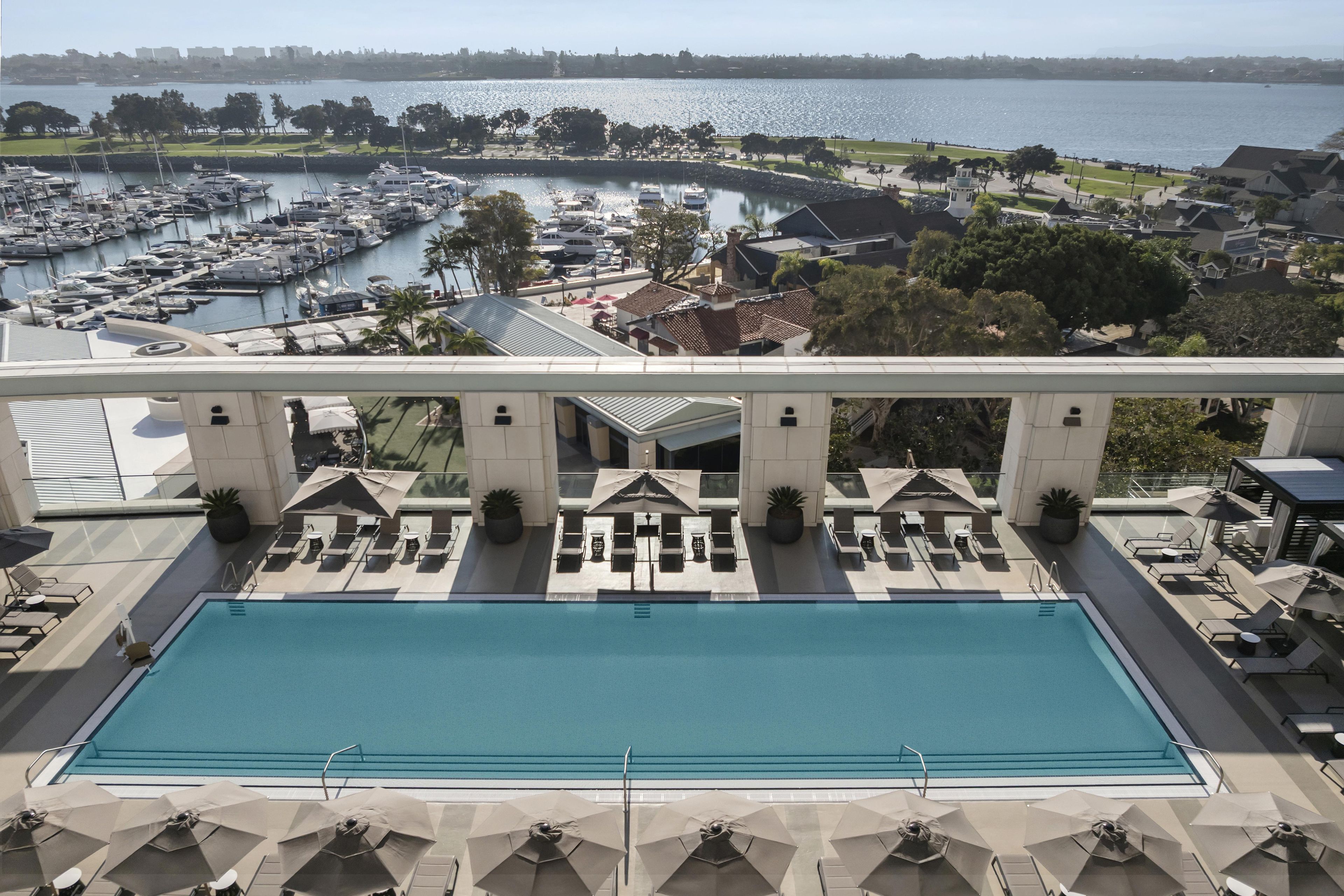 A pool with lounge chairs and umbrellas, a marina with boats, and a view of a cityscape across the water.