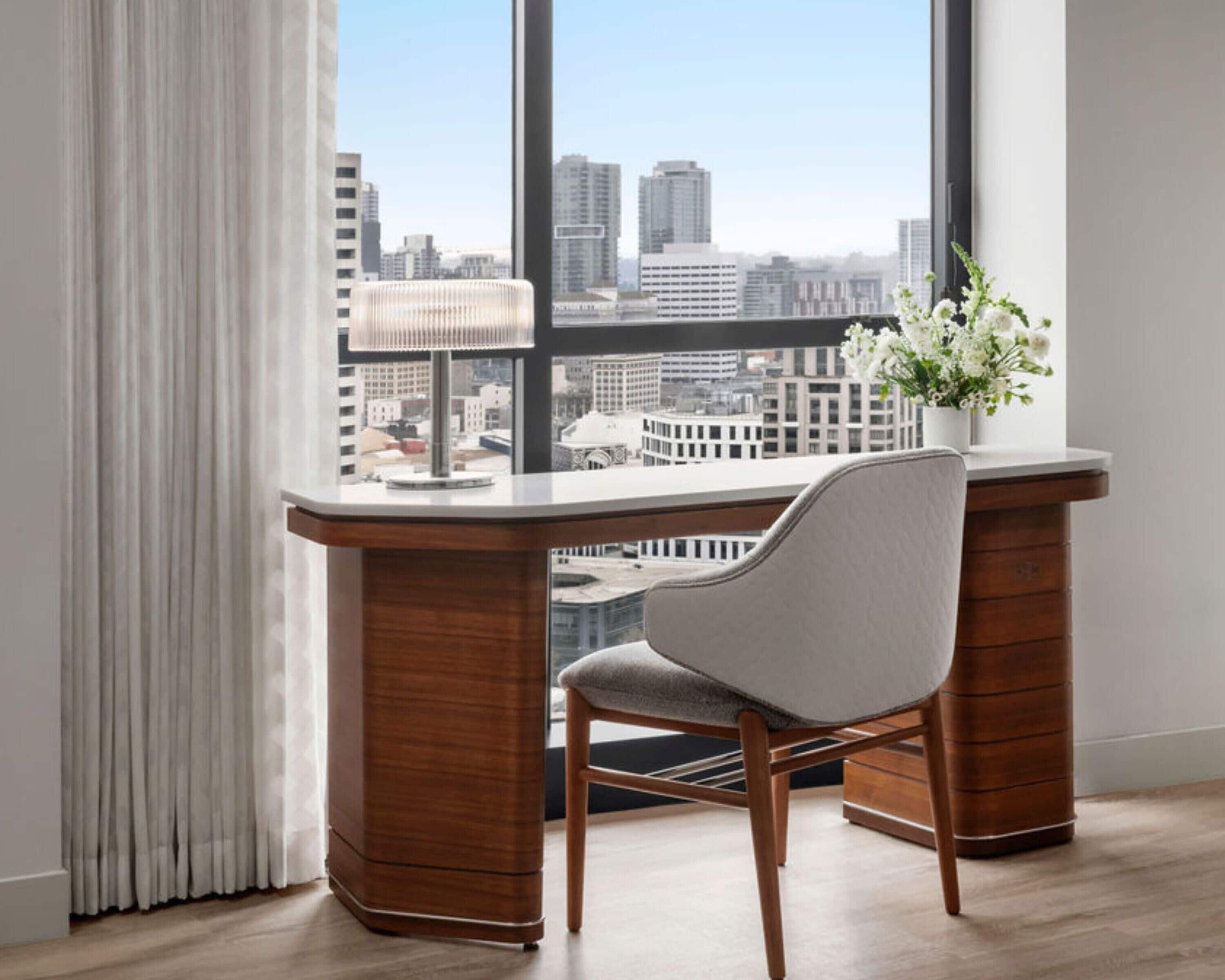A modern office desk with a chair, a lamp, and a vase with flowers, overlooking a cityscape through large windows.