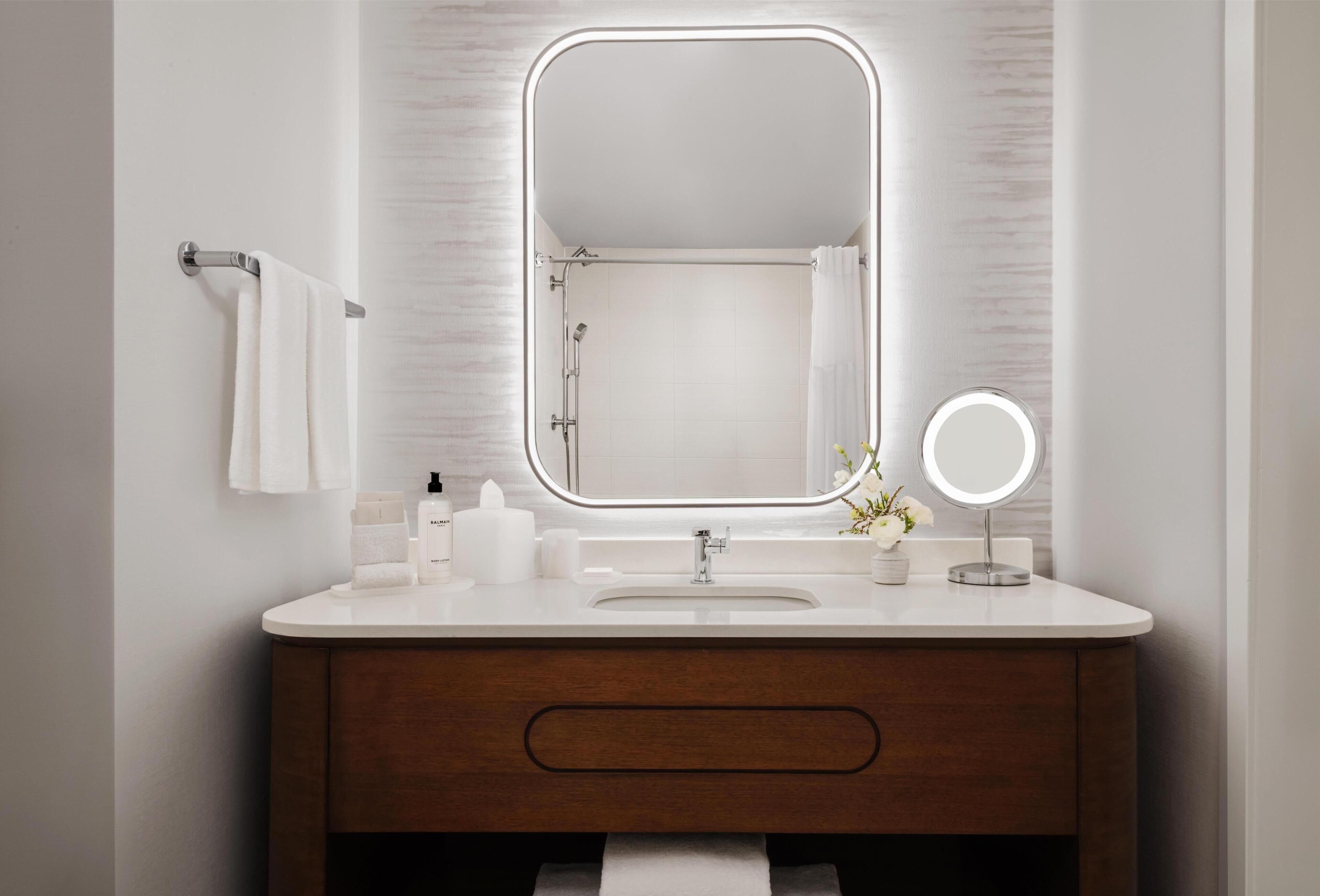 A bathroom with a white vanity, a large mirror with a light, a towel rack, and a small round mirror.