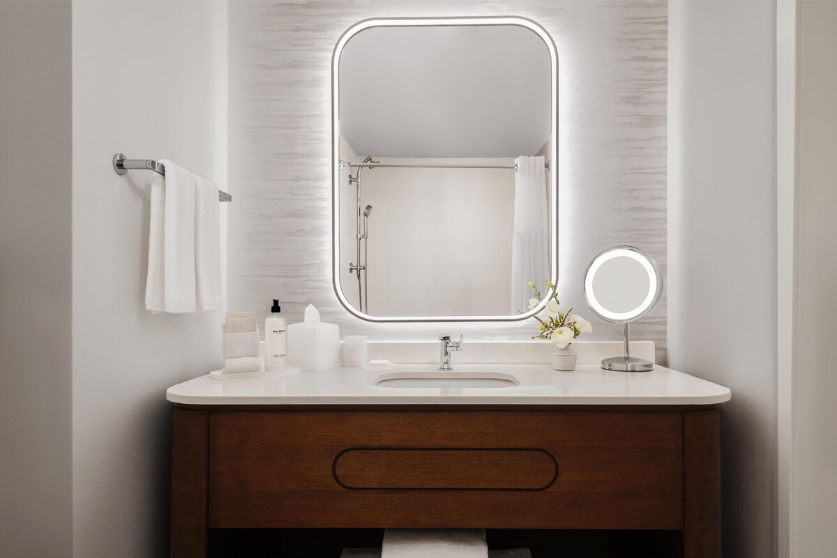 Une salle de bain avec un meuble-lavabo blanc, un grand miroir avec un éclairage intégré, un porte-serviettes et un petit miroir rond.