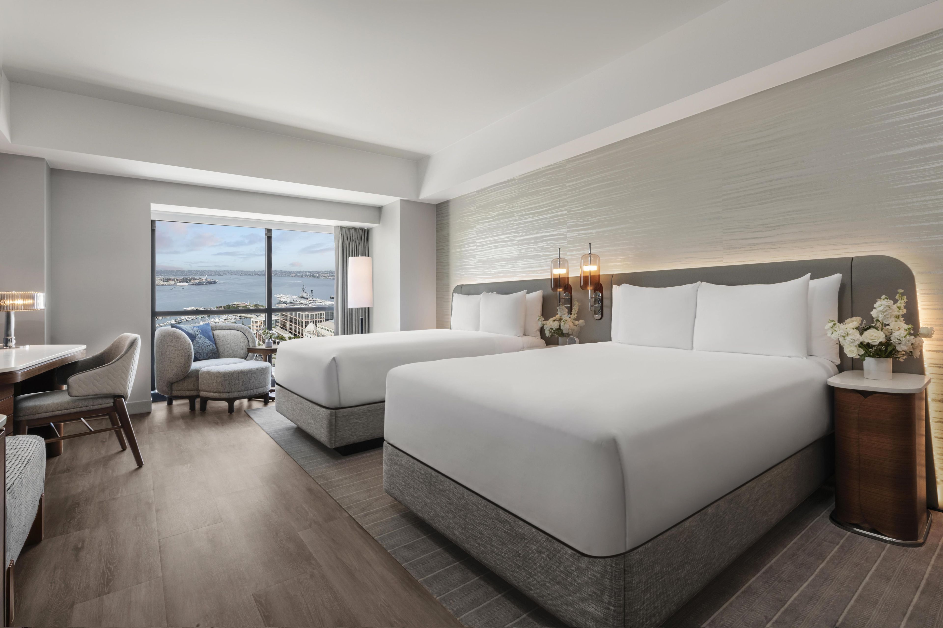 A hotel room with two beds, a desk, a chair, and a view of the water.