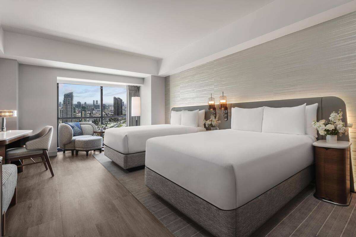 A hotel room with two beds, a desk, a chair, and a view of the cityscape.
