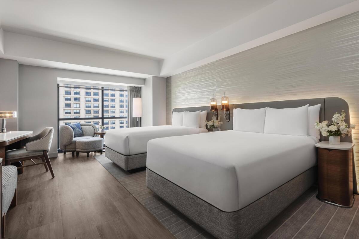 A hotel room with two beds, a desk, a chair, and a view of a building through a large window.