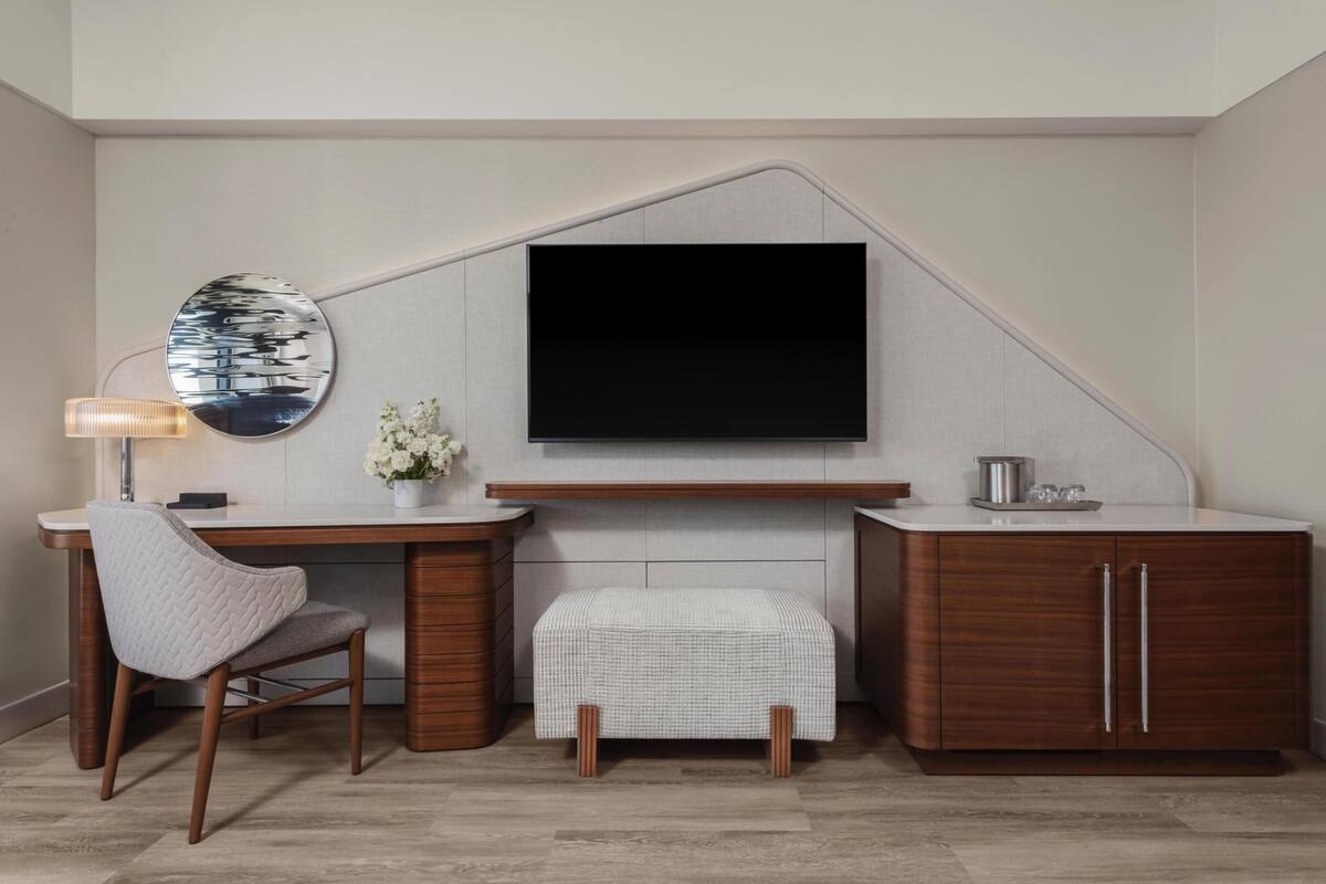 A modern room with a flat-screen TV mounted on a wall, a wooden desk with a chair, a small white ottoman, and a cabinet with drawers.
