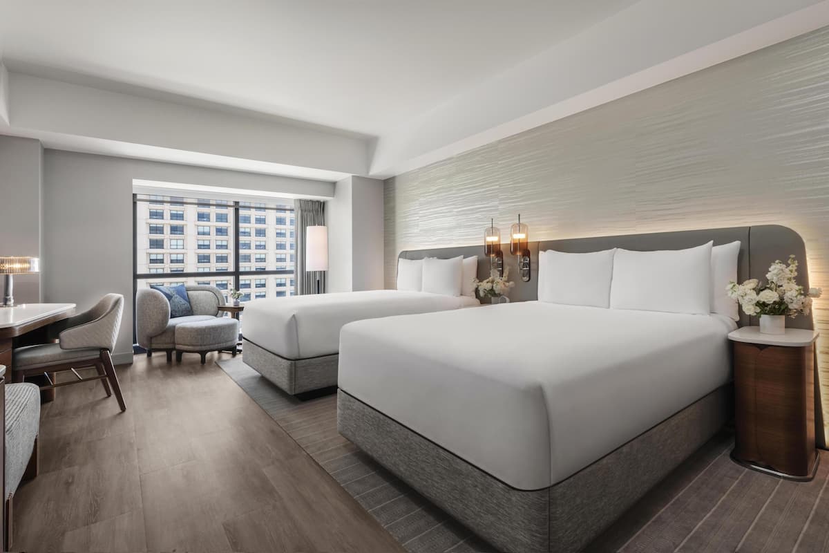 A hotel room with two beds, a desk, a chair, and a view of a building through a large window.