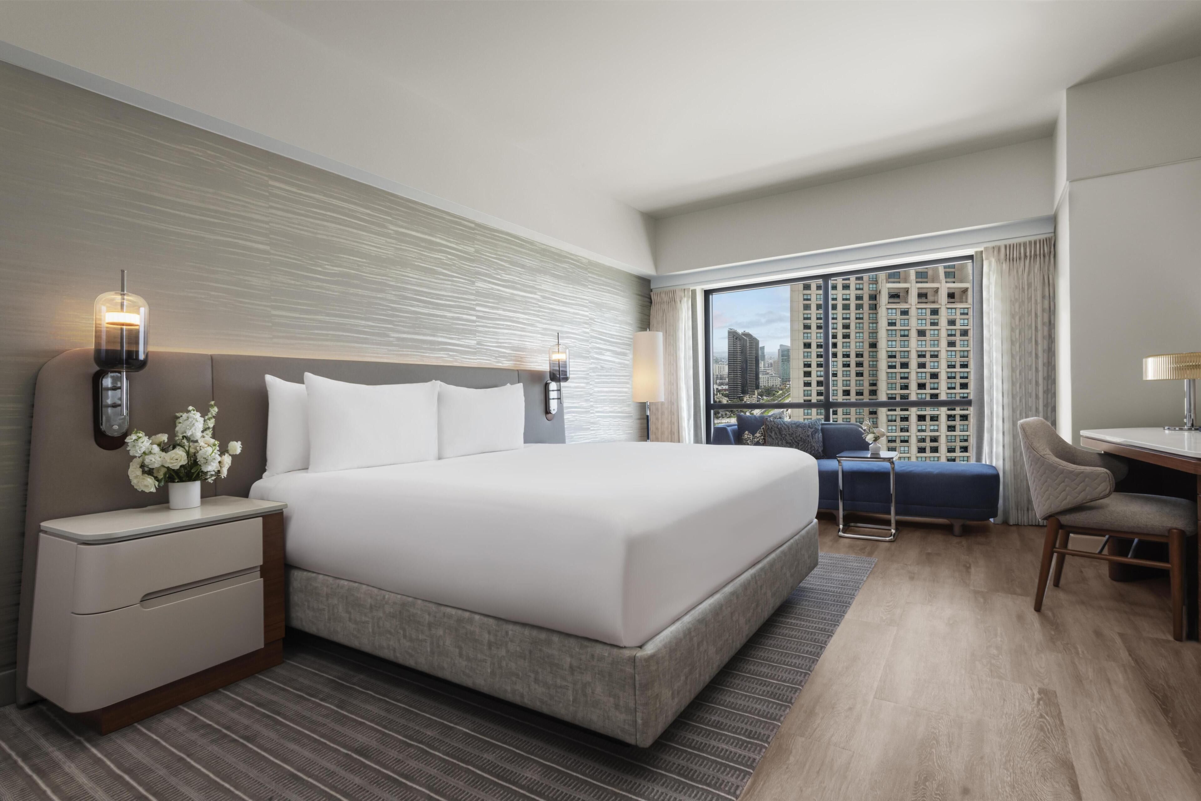 A modern hotel room with a large bed, bedside tables, a desk, and a view of a cityscape through large windows.