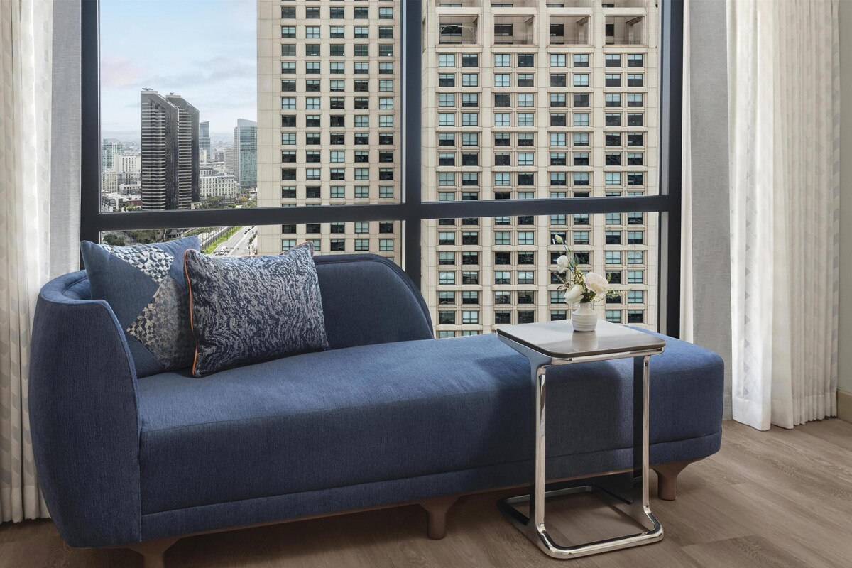 A modern living room with a blue sofa, a small side table, and a view of high-rise buildings through large windows.