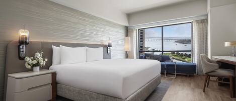 A modern hotel room with a large bed, a desk, and a view of a bridge and water.