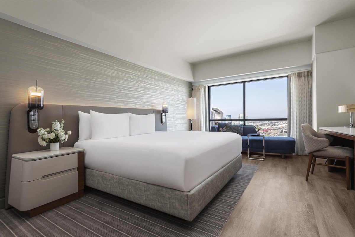A modern hotel room with a large bed, a desk, and a view of the city.
