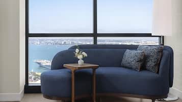 A room with a blue sofa, a small wooden side table with a vase of flowers, and a view of a body of water and distant buildings.