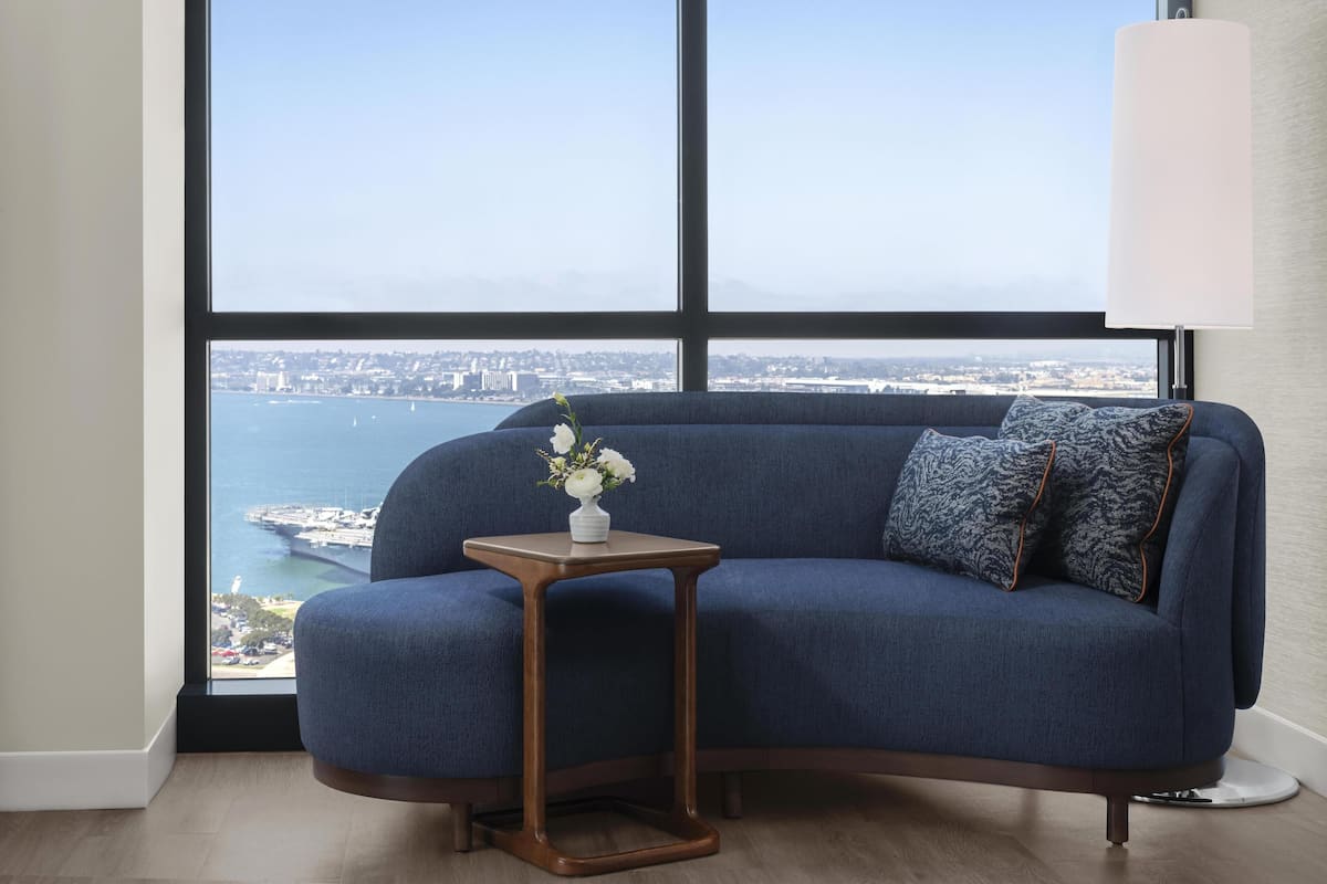 Une pièce avec un canapé bleu, une petite table d’appoint en bois sur laquelle est posé un vase de fleurs, et une vue sur une étendue d’eau et des bâtiments éloignés.