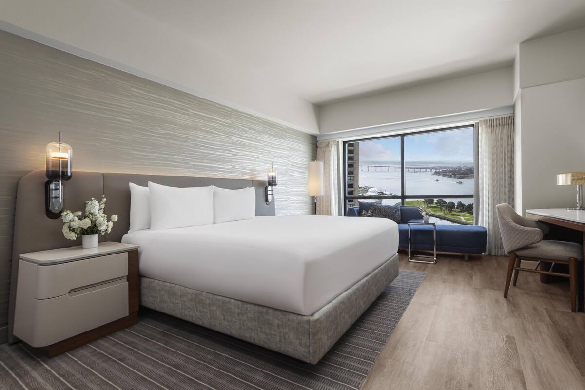 A modern hotel room with a large bed, a desk, and a view of a bridge and water.