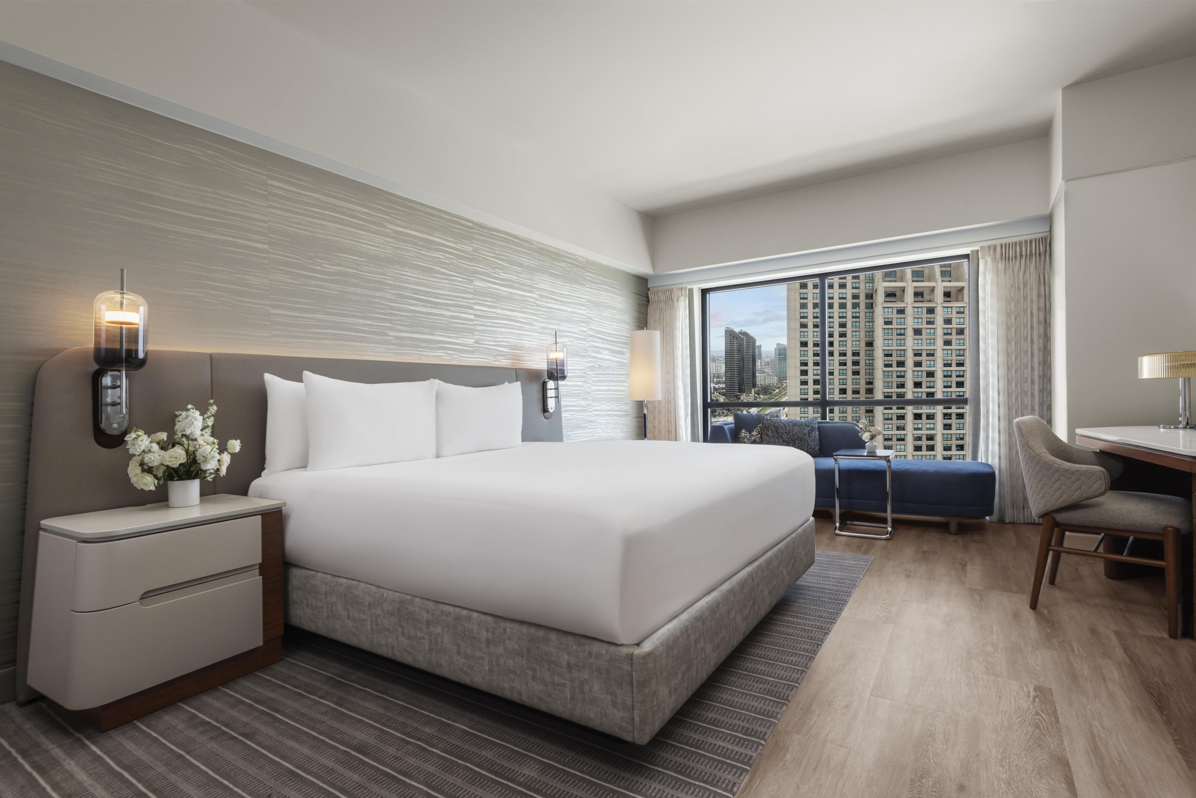Une chambre d’hôtel moderne dotée d’un grand lit, de tables de chevet, d’un bureau et offrant une vue sur le paysage urbain grâce à de grandes fenêtres.