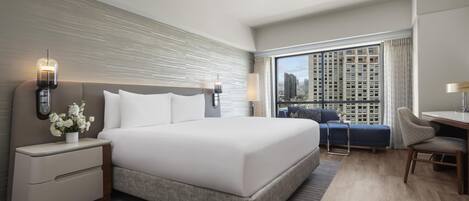 Une chambre d’hôtel moderne dotée d’un grand lit, de tables de chevet, d’un bureau et offrant une vue sur le paysage urbain grâce à de grandes fenêtres.